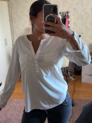Vit långärmad blus med fickor - Stilren vit blus med långa ärmar och två bröstfickor. Blusen har en avslappnad passform och en rundad halsringning med liten v-ringad öppning framtill. Perfekt att matcha med jeans eller kjol för en enkel och snygg look. Säljer för 100kr. 