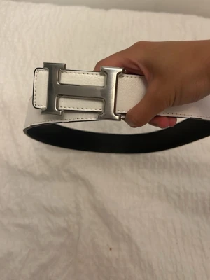 Vitt skärp med stor silverspänne - Snyggt vitt skärp med ett stort H-format silverspänne. Bältet är gjort i slätt skinn och har flera hål för justering. Perfekt accessoar för att lyfta din outfit och ge en clean look.
