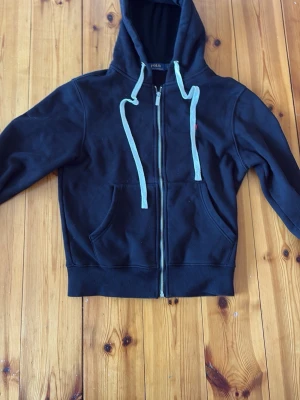 Svart hoodie med dragkedja och huva - Säljer en svart hoodie med dragkedja framtill och stora fickor. Tröjan har långa ärmar, ribbade muddar och en huva med vita snören. Perfekt för en avslappnad stil och enkel att matcha med jeans eller joggers.