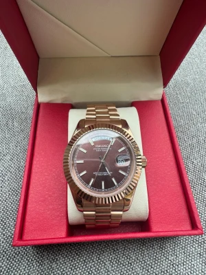 Seiko klocka i roséguld med länk - Säljer en stilren Seiko klocka i roséguld med länkarmband och mörkröd urtavla. Klockan har dag- och datumvisning samt markerade index. Kommer i en snygg röd box, perfekt för dig som vill ha en elegant accessoar.