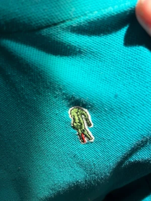 Turkos pikétröja från Lacoste - Snygg turkos pikétröja från Lacoste med klassisk krage och korta ärmar. Tröjan har den ikoniska krokodilloggan broderad på bröstet och är tillverkad i ett mjukt pikématerial. Perfekt för en avslappnad och sportig stil.