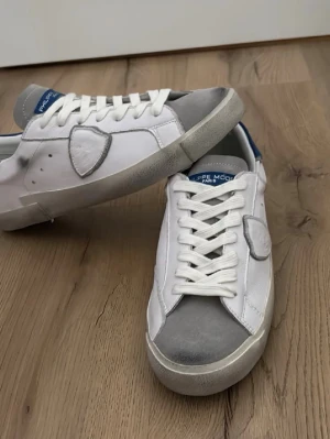 Vita Philippe Model sneakers med grå detaljer - Snygga vita sneakers från Philippe Model med grå mockadetaljer på tå och häl samt blå hälflik. Klassisk låg modell med vita skosnören och ikonisk sköld på sidan. Perfekta för en clean och trendig look.