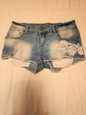 Blå jeansshorts med spetsdetaljer - Blå jeansshorts med slitna detaljer och rå kant. Låg midja. Dekorerade med vita spetsblommor och pärlor på ena sidan framtill. Klassisk femficksmodell med knapp och dragkedja.  Vintage, size S, midjemått 32,5cm