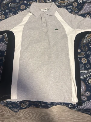 Grå pikétröja från Lacoste - Säljer en grå pikétröja från Lacoste med vit och svart panel på sidorna. Tröjan har klassisk krage, knappar framtill och den ikoniska gröna krokodilloggan på bröstet. Relaxed fit och tillverkad i mjuk bomull. Perfekt för en clean och sportig look.