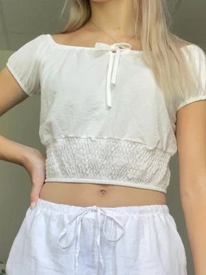 Vit kortärmad blus från Chiquelle - Vit kortärmad blus från Chiquelle i storlek xs. Använd 2-3 gånger. Går att justera med snörningen som önskas och den går att ha off shoulder om man vill då den är stretchig i urringningen. Smockad del på magen av tröjan. Normal i storleken och passar xs-s. Lite genomskinlig men i den mån som vita tröjor brukar vara