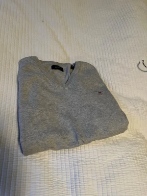 Ljusgrå V-ringad tröja från GANT - Klassisk ljusgrå V-ringad tröja från GANT med liten broderad logga på bröstet. Tröjan är i mjukt material och har ribbade muddar vid hals, ärmslut och nederkant. Perfekt för en stilren och enkel look.