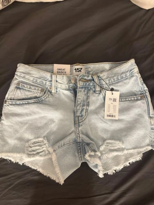 Ljusblå denimshorts med slitningar - Säljer ett par ljusblå denimshorts från 157 med råa kanter och slitna detaljer framtill. Shortsen har fem fickor, bälteshällor och klassisk knappgylf. Perfekta för varma dagar och ger en avslappnad vibe.