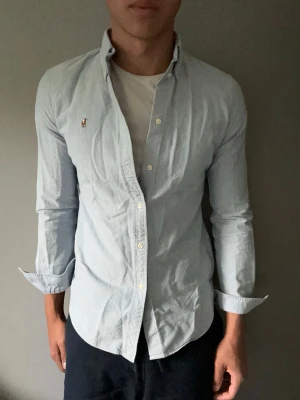 Ljusblå skjorta med knappdetaljer - Snygg ljusblå skjorta med klassisk krage och knappar framtill. Skjortan har lång ärm och en liten broderad logga på bröstet. Perfekt för en clean och stilren look. Tillverkad i mjuk bomull för skön känsla.
