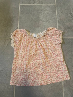 Blommig topp från Lindex Kids - Söt blommig topp från Lindex Kids i storlek 116. Toppen har korta ärmar, volangdetaljer vid halsringningen och ett rosa och beige blommönster på ljus botten. Perfekt för vår och sommar!