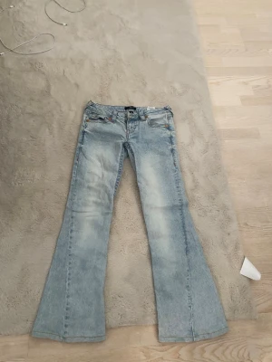 Ljusblå bootcut jeans med låg midja - Säljer ett par ljusblå bootcut jeans med låg midja och klassiska fem fickor. Jeansen har slitna detaljer och är tillverkade i denim med kontrastsömmar. Perfekta för dig som gillar retrovibbar och en avslappnad look.