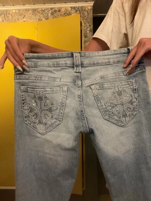 Ljusblå jeans med broderade fickor Ny - Säljer ett par ljusblå jeans med snygga broderade detaljer och små stenar på bakfickorna. Jeansen har rak passform, klassisk femficksdesign och bälteshällor. Perfekta för dig som vill ha något unikt och trendigt i garderoben.