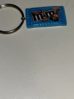 Nyckelring med en blå M&M - Säljer en rolig nyckelring med en liten blå    M&M's-förpackning. Perfekt att hänga på väskan eller nycklarna för en färgglad touch. Tillverkad i plast och metall.
