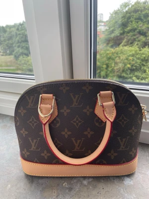  handväska - Snygg handväska från i klassiskt brunt monogrammönster med beige och röda detaljer. Väskan har rundad form, två handtag och guldfärgade metalldetaljer. Tillverkad i canvas med läderdetaljer. Perfekt för dig som gillar lyxig och tidlös design. band tillkommer