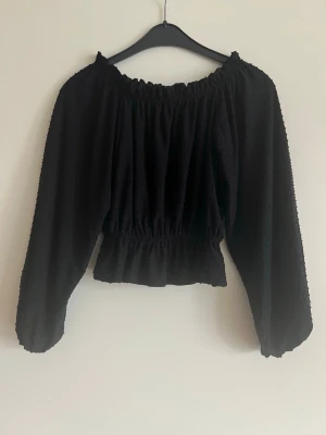 Svart offshoulder blus med struktur - Trendig svart offshoulder blus med långärmad design och resår vid axlar och midja. Blusen har ett diskret prickigt strukturmönster i tyget och volang nedtill. Perfekt för dig som gillar en avslappnad men ändå chic stil. Längd ca 44cm. 