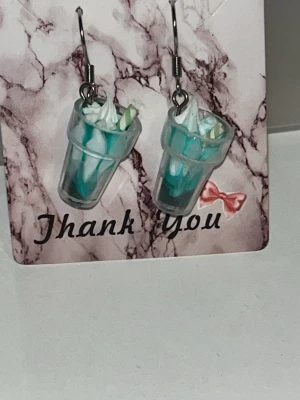 Örhängen med milkshake-design - Unika örhängen formade som små milkshakes i plast med turkosa och vita detaljer samt sugrör. Örhängens krokarna är i rostfritt stål. Finns i flera färger som rosa/blå/grå/brun.