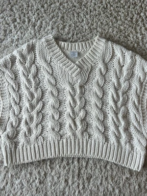 Beige kabelstickad cropped väst - Säljer en beige kabelstickad väst i croppad modell med V-ringning och bred ribbad kant. Västen har ett chunky flätmönster framtill och är gjord i mjukt garn, perfekt att styla över en skjorta, en tunn tröja eller t-shirt.