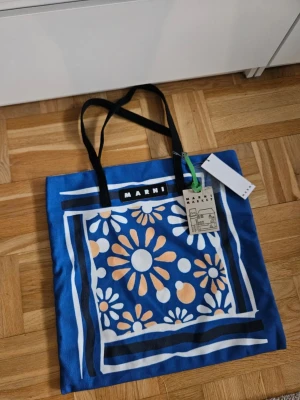 Marni shoppingväska med blommor från Marni - Snygg blå toteväska från Marni med vita och orange blommor i ett grafiskt mönster. Väskan har svarta handtag och en tydlig Marni-logga framtill. Perfekt för att bära allt du behöver och ger en färgklick till din outfit.