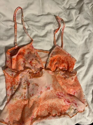 Blommigt orange linne med volang - Säljer ett sött loungewear-linne i tunn, luftig mesh med blommigt mönster i orange, rosa och vitt. Linnet har smala axelband och volangkant nedtill, perfekt för chill dagar hemma. Materialet känns lätt och flowy, och toppen har en feminin vibe.