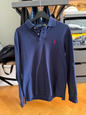 Marinblå långärmad piké från Polo Ralph Lauren - Klassisk marinblå långärmad pikétröja från Polo Ralph Lauren med krage, knäppning vid halsen och den ikoniska röda logotypen broderad på bröstet. Tillverkad i mjuk bomull som ger en avslappnad men ändå stilren look.