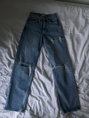 Ljusblå raka ripped jeans - Säljer ett par ljusblå jeans med raka ben och hög midja. Jeansen har slitningar och hål vid knäna och låren för en trendig, avslappnad look. Klassisk femficksmodell i denim med råa kanter vid bensluten.