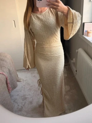Lång beige klänning med vida ärmar - Trendig långklänning i beige med strukturerat tyg och vida, långa ärmar. Klänningen har en kroppsnära passform och rund halsringning. Perfekt för dig som gillar en stilren och elegant look. Schruch med knytning på sidan 