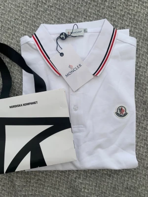 Vit Moncler pikétröja med röd detalj - Klassisk vit pikétröja från Moncler med krage och knappar framtill. Kragen har snygga röda och svarta ränder och bröstet pryds av Monclers ikoniska logotyp. Perfekt till jeans eller shorts för en clean och stilren look.