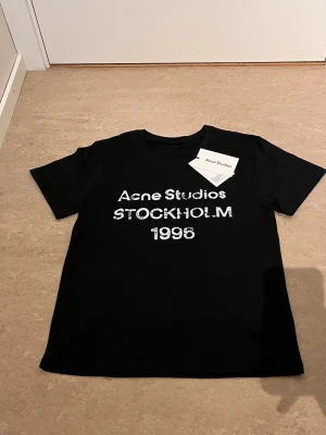 Svart Acne Studios t-shirt - Snygg svart t-shirt från Acne Studios med vit text på bröstet där det står 'Acne Studios STOCKHOLM 1996'. Klassisk passform och rund hals. Perfekt för dig som gillar stilrena plagg med en twist.