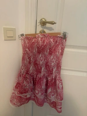 rosa och vit kortklänning med volang - Supersöt kortklänning i rosa och vitt med paisleymönster och volangkant. Klänningen är axelbandslös och har smock upptill för en bekväm passform. Perfekt för varma dagar och sommarens alla äventyr.