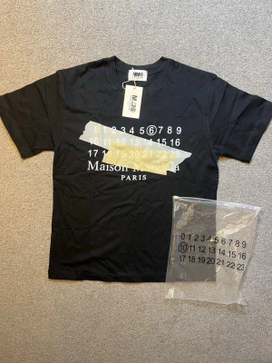 Svart Maison Margiela t-shirt - Cool svart t-shirt från Maison Margiela med tryck av siffror och logga på bröstet. T-shirten har korta ärmar och rund hals. Materialet känns mjukt och bekvämt, perfekt för en trendig vardagslook.