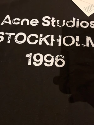 Svart Acne Studios t-shirt Stockholm - Snygg svart t-shirt från Acne Studios med tryck i vitt framtill där det står 'Acne Studios STOCKHOLM 1996'. Klassisk passform och rund hals. Perfekt för dig som gillar stilrena och trendiga plagg.