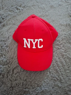 Röd keps med NYC-brodyr - Snygg röd keps med vit brodyr där det står 'NYC' framtill. Klassisk böjd skärm och justerbar baksida. Perfekt för dig som vill ha en streetstyle-look och gillar enkla, tydliga detaljer. Materialet känns som bomull.