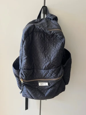 Svart quiltad ryggsäck från DAY ET - Snygg svart quiltad ryggsäck från DAY ET med guldfärgade dragkedjor och flera fack. Rymlig modell med två sidofickor och ett stort huvudfack. Tillverkad i vadderat tyg med mönstrad struktur och klassisk DAY ET-logga framtill.