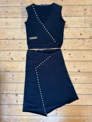 Vintage Rodebjer år 2000; Svart set i bomull med nitar  - Vintage Rodebjer år 2000! Första kollektionen! Inköpt i liten butik hörnet Birger Jarlsgatan/ Riddargatan. Snyggt svart tvådelat set med topp och kjol i bomull/ ”collegetyg”. Toppen har V-ringning och dekorativa bronsfärgade nitar längs omlottkanten samt en liten patchdetalj. Kjolen har asymmetrisk nederkant och matchande nitar. Perfekt skick! Top storlek S, kjol storlek M. Passar S och XS