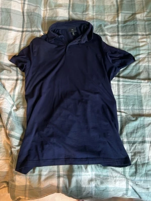 Mörkblå pikétröja med krage - En klassisk mörkblå pikétröja med krage och korta ärmar. Tröjan har knappar vid halsen och är tillverkad i ett mjukt material som känns bekvämt mot huden. Perfekt för en avslappnad men ändå stilren look.