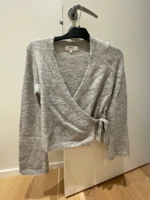Grå omlottkofta med knytband - Mysig grå omlottkofta i mjukt stickat material med långa ärmar och knytband i midjan. Perfekt för lager på lager och enkel att styla till olika outfits. Koftan har en avslappnad passform och ribbade kanter.