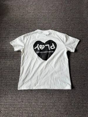 T-shirt - Vit t-shirt med svart hjärta och texten 'PLAY fantasy and miracle' på ryggen samt ett mindre hjärta med text på bröstet. Klassisk rund hals och korta ärmar. Perfekt för dig som gillar grafiska tryck och enkel stil.
