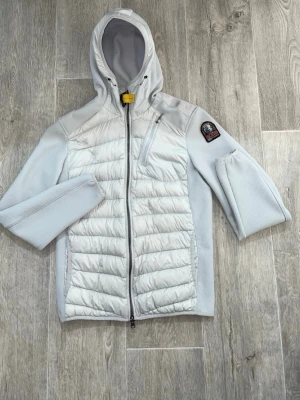 Vit cardigan från Parajumpers - Snygg vit pufferjacka från Parajumpers med huva och dragkedja framtill. Jackan har quiltade partier på framsidan, mesh-liknande tyg på ärmar och sidor samt en bröstficka med dragkedja. Klassisk Parajumpers-logga på ärmen och gul detalj vid halsen. Skickar samma dag vid köp, storlek S passar M