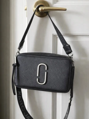 Svart axelväska Marc Jacobs - Säljer en svart axelväska från Marc Jacobs med silverfärgad logga framtill. Den är i mycket bra kvalite. Väskan har justerbar axelrem med logotyp och dragkedja upptill. Stilren och praktisk modell i ett tåligt material som passar till många outfits. Aldrig använd. Den är lite ihoptryckt ovanpå men det är för att den inte har använts. Ej äkta. 