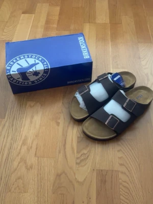  Birkenstock  - Säljer ett par klassiska Birkenstock sandaler i brunt med två breda remmar och justerbara metallspännen. Fotbädden är i mocka och sulan är ergonomiskt utformad för extra komfort. Perfekta för dig som gillar stilren och avslappnad look. Pris kan diskuteras 