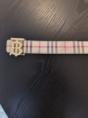 Burberry bälte  - Snyggt vändbart bälte med klassiskt rutigt mönster på ena sidan i beige, rosa och grå toner och svart på andra sidan. Bältet har ett stilrent guldspänne med initialer. Perfekt accessoar för att lyfta din outfit.