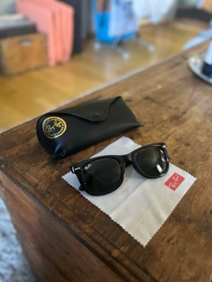 Svarta Ray-Ban Wayfarer solglasögon - Klassiska svarta Ray-Ban Wayfarer solglasögon med glansig båge och mörka linser. Kommer med original Ray-Ban fodral i svart skinn och putsduk. Perfekt accessoar för dig som vill ha en tidlös och ikonisk look.