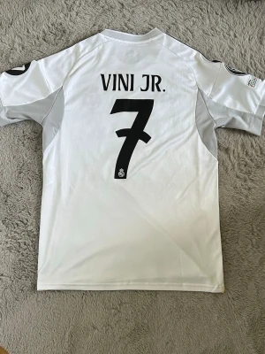 Real Madrid 25/26 Vini Jr fotbollströja - Säljer en vit Real Madrid matchtröja från Adidas med svarta detaljer och gula linjer. Tröjan har klubbmärke på bröstet, svart Adidas-logga och sponsortryck. Tillverkad i lätt och ventilerande material, perfekt för fotboll eller träning.