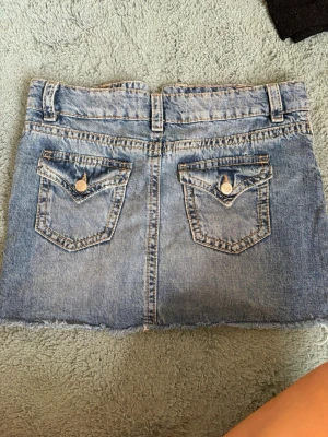 Ljusblå jeanskjol med fickor - Kort jeanskjol i ljusblå denim med två bakfickor med lock och knappdetaljer. Kjolen har rå kant nedtill och bälteshällor i midjan. Perfekt för en avslappnad och trendig look.
