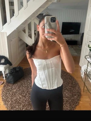 Vit axelbandslös satin-korsett - Snygg vit korsett i satin med axelbandslös design och markerade sömmar framtill för en klassisk siluett. Materialet har en lätt glans och ger en elegant look. Perfekt att styla med jeans eller kjol för en trendig outfit.