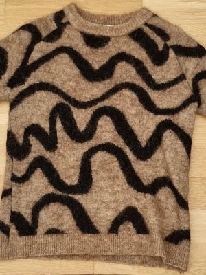 Beige stickad tröja Carin Wester - Stickad tröja från Carin Wester i beige med svarta vågiga mönster. Tröjan har korta ärmar, rund halsringning och är gjord i ett fluffigt, mjukt material som ger en mysig känsla. Perfekt för dig som gillar unika stickade plagg.