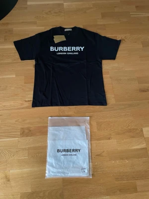 Svart Burberry t-shirt med logga - Snygg svart t-shirt från Burberry med stor vit logga och texten 'London England' på bröstet. Klassisk rund halsringning och korta ärmar. Tillverkad i mjuk bomull för en skön känsla. Perfekt för dig som gillar stilrena och ikoniska plagg. Står storlek S men är storlek M 