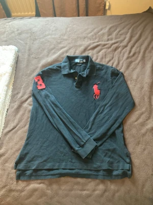 Svar långärmad piké från Ralph Lauren - Säljer en svart långärmad pikétröja från Ralph Lauren med röd broderad logga på bröstet och rött nummer på ärmen. Klassisk krage och knappar vid halsen. Perfekt för dig som gillar stilren och sportig look.