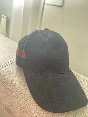 Svart keps med grön-röd rand - Snygg svart keps med diskret mönster och en grön-röd rand på sidan. Klassisk böjd skärm och justerbar passform. Materialet ser ut att vara bomull eller bomullsblandning. Perfekt för dig som vill ha en stilren accessoar med en touch av färg.