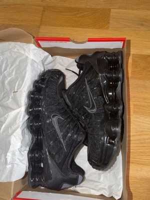 Nike Shox svarta sneakers - Nike Shox sneakers i helsvart med ikoniska fjädrande sulor och meshdetaljer. Skorna har en sportig look med silvriga Swoosh-loggor på sidorna och röda detaljer på plösen. Tillverkade i en mix av syntetmaterial och mesh för bra andningsförmåga. Helt nya, använda en gång.