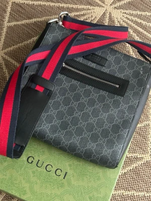 Gucci axelväska med GG-mönster - Snygg axelväska från Gucci i mörkgrått med klassiskt GG-mönster. Väskan har en svart dragkedjeficka framtill och justerbar axelrem i rött och marinblått. Tillverkad i canvas med läderdetaljer. Perfekt för dig som vill ha en stilren och trendig accessoar.